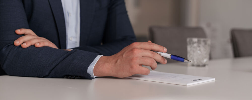 Detailaufnahme am Besprechungstisch: Eine Person im Anzug hält einen Stift über einem Notizblock – als Symbol für die drei Schritte zur Zusammenarbeit und die nächsten konkreten To-dos.
